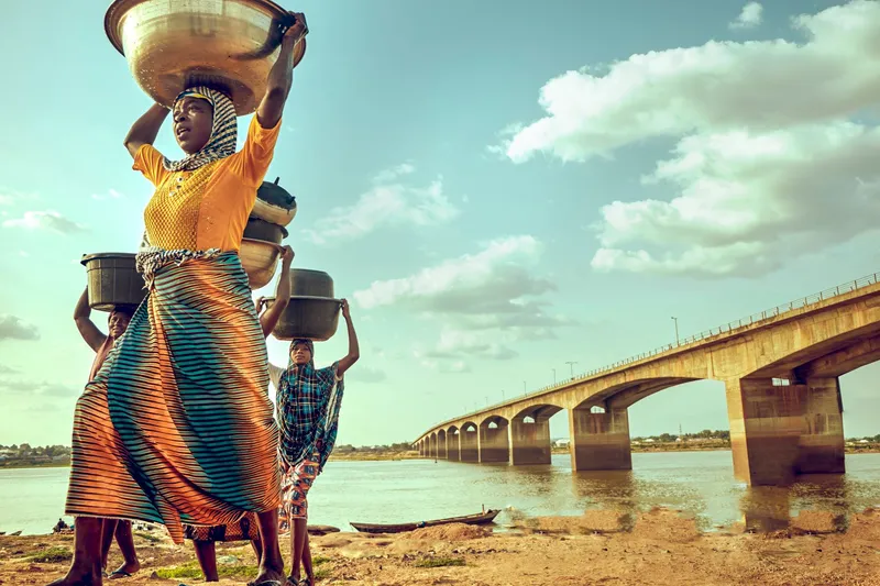 Women carrying bowls along the bank of the Benue River, with the Makurdi bridge in the background