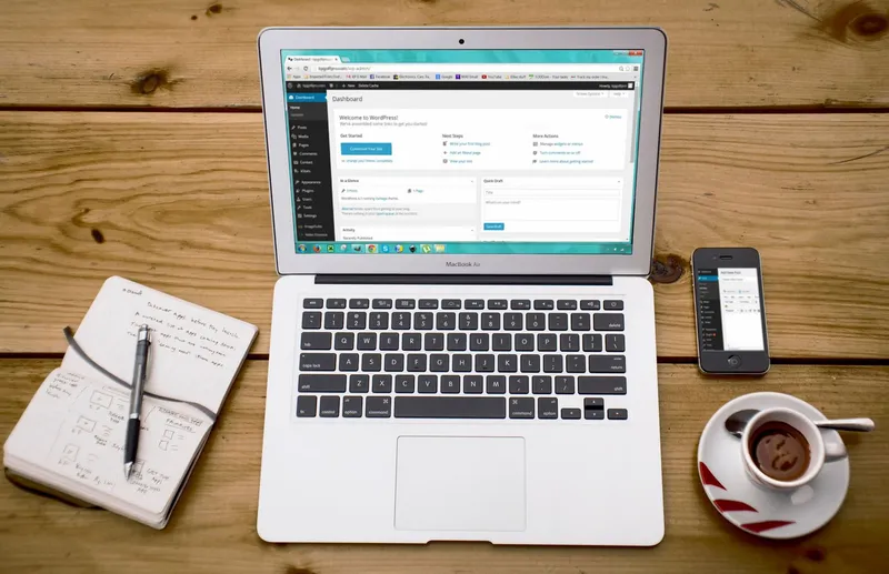 A MacBook Air open beside a smartphone, cup of tea, and notebook on a wooden desk